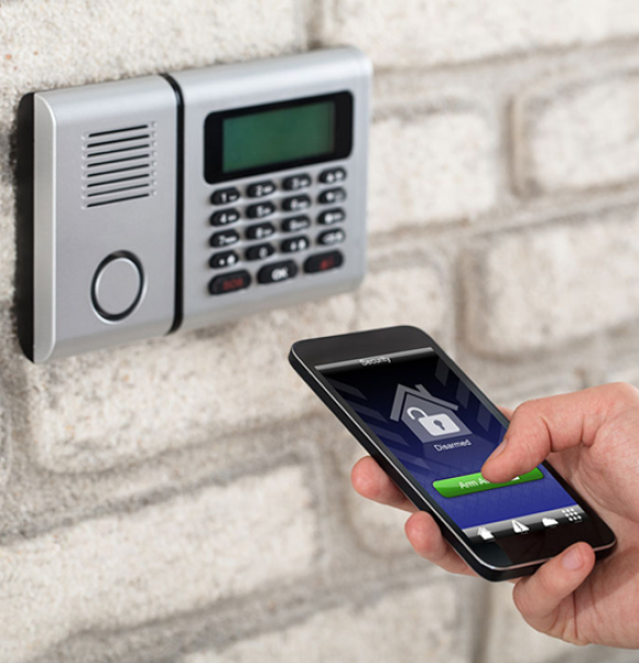 Close-up of a person's hand using a smartphone to arm a home security system via a dedicated app, with the security keypad mounted on a white brick wall in the background.