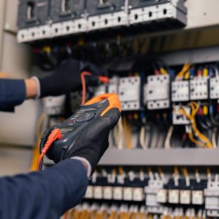 A close-up view of a technician's hands wearing black gloves and holding a multimeter with orange and red probes to test wires inside a complex electrical control panel.
