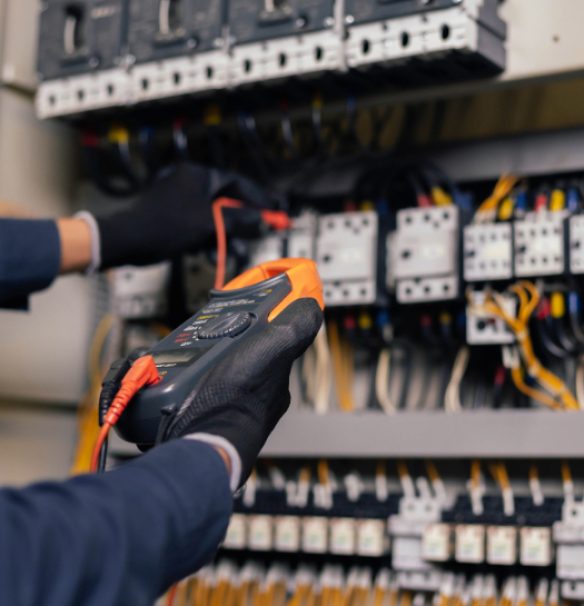 A close-up view of a technician's hands wearing black gloves and holding a multimeter with orange and red probes to test wires inside a complex electrical control panel.