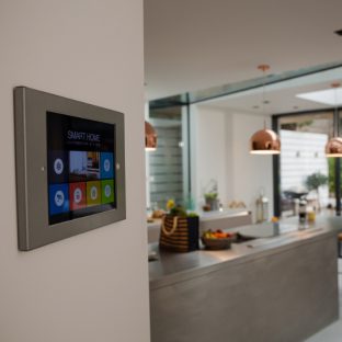 A wall-mounted touchscreen device displaying a smart home automation interface with various icons, situated in a modern, open-plan kitchen.